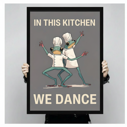 Person holding a large black framed funny frog chef kitchen dance canvas art print showing print size scale with two green frogs in white chef uniforms dancing and the humorous quote In This Kitchen We Dance on a grey background