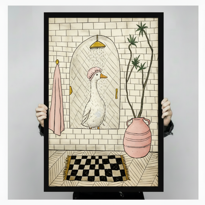 Person holding a black framed funny goose in shower cap bathroom art print showing full illustration detail with gold showerhead, pink towel, checkerboard rug, and palm plant against a light gray backdrop
