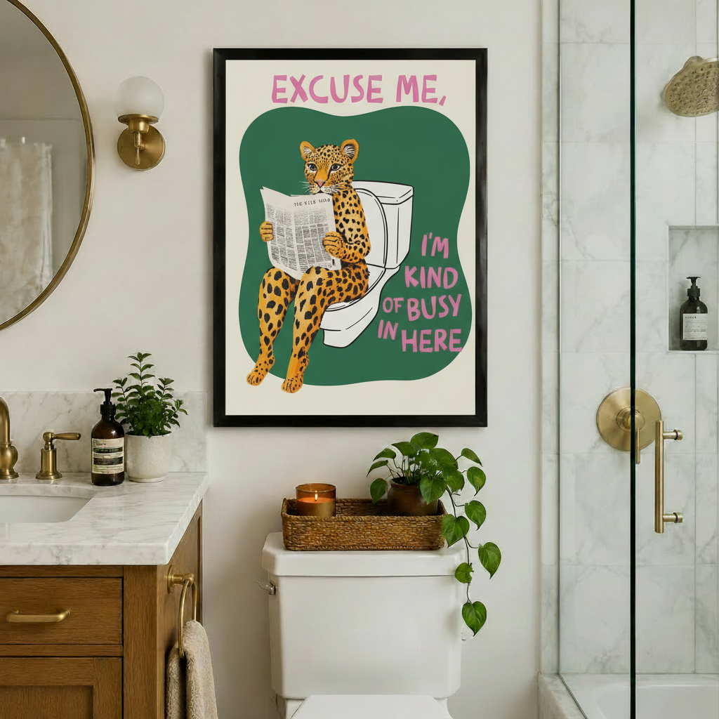 Humorous leopard bathroom wall art in a black frame with pink and green "Excuse Me I'm Kind of Busy In Here" text, hung above a toilet in a luxury bathroom with marble countertops, gold fixtures, and trailing ivy