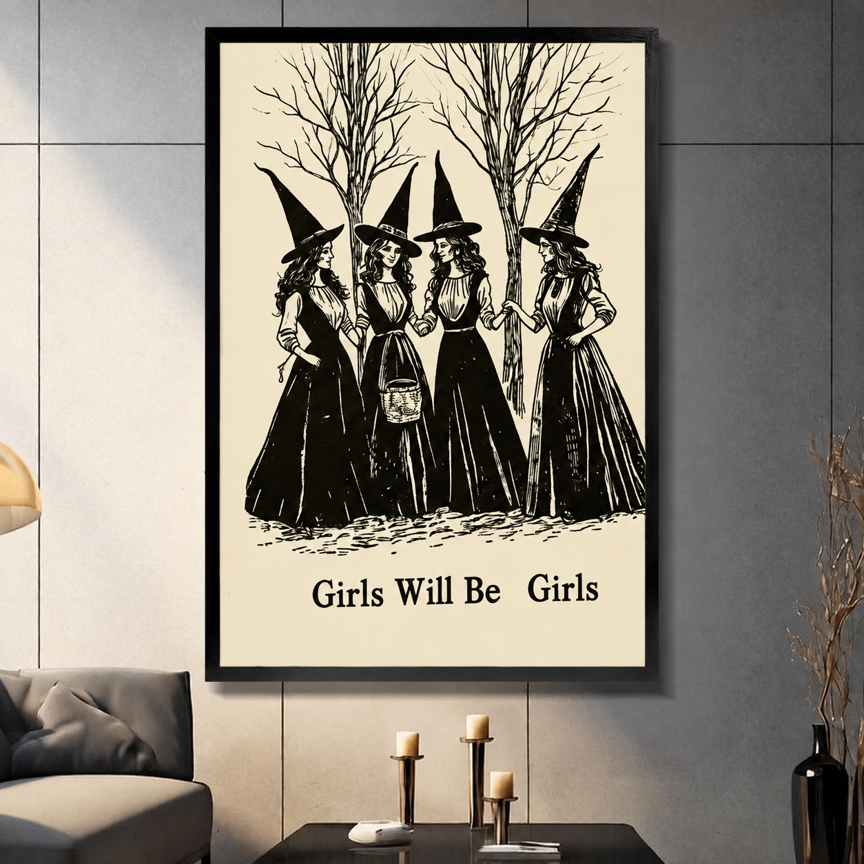 Framed gothic wall art print of four witches in black dresses with pointed hats standing among bare trees, titled Girls Will Be Girls, displayed in a modern living room above a dark coffee table with candles