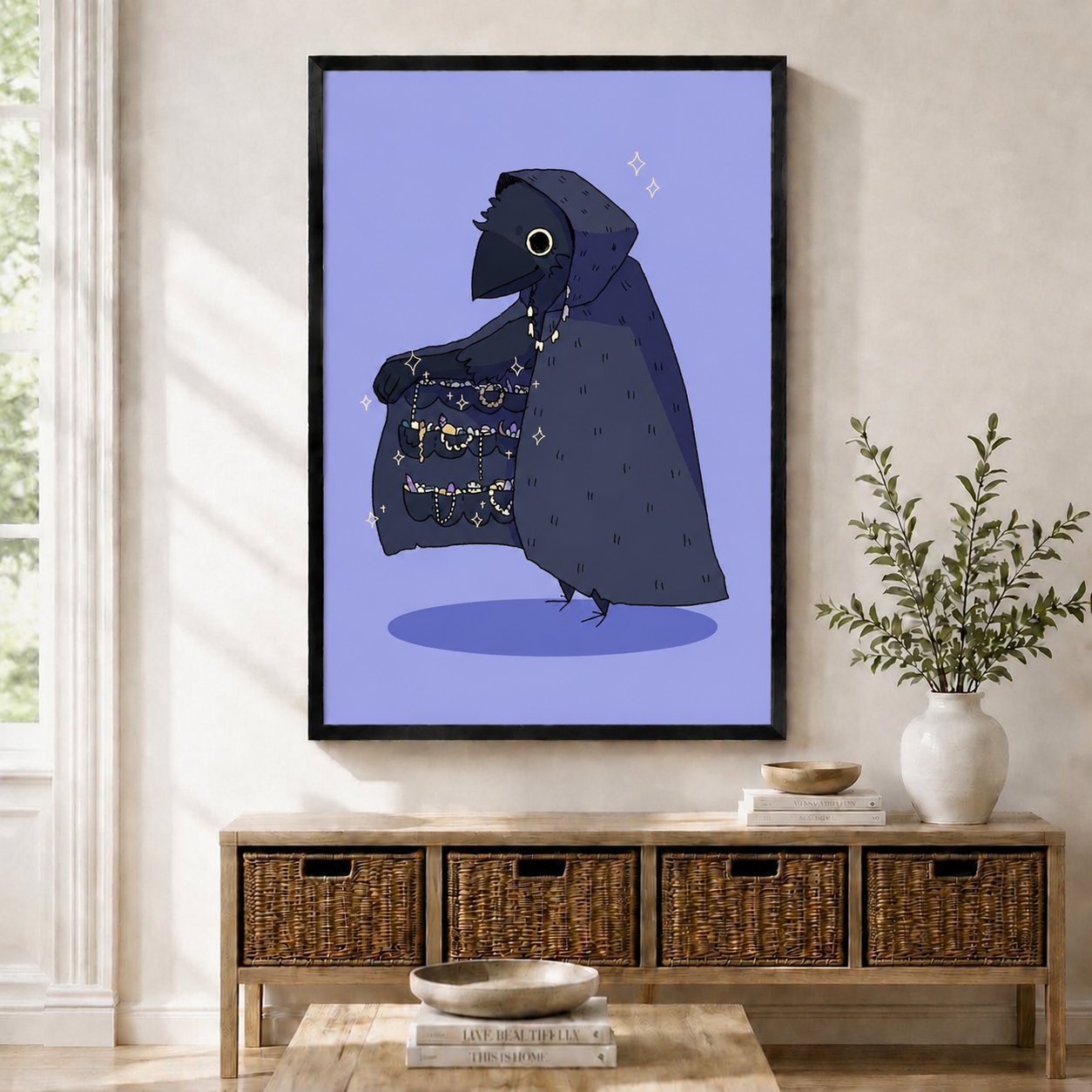 Black-framed gothic crow wall art print featuring a hooded raven displaying a sparkling treasure cloak, hung above a wicker storage console in a Scandinavian entryway
