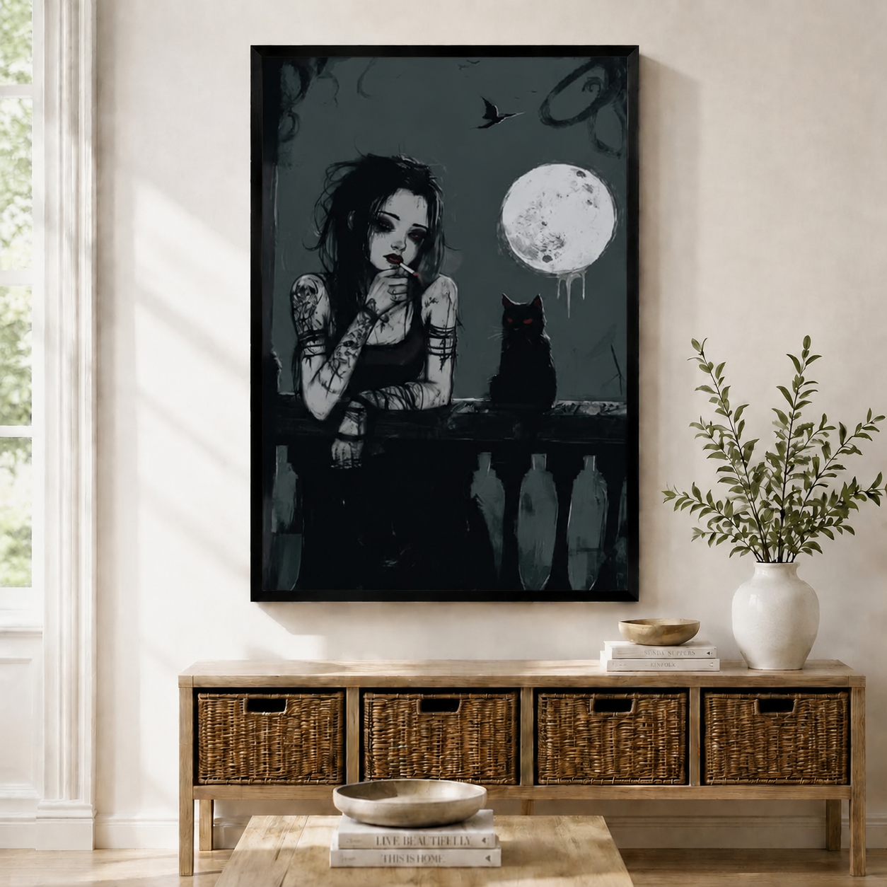 Framed dark gothic wall art print of a tattooed girl leaning on a balcony railing beside a black cat under a glowing full moon at night, displayed above a wicker storage console in a bright Scandinavian living room