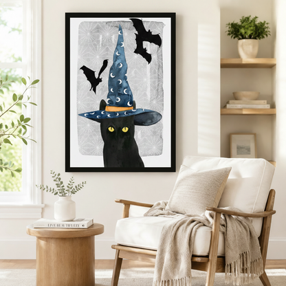 Black framed Halloween witch black cat with bat canvas art showing a stealthy black cat with vivid yellow eyes under a wide-brimmed blue moon-print witch hat with black bats flying above on a crackled misty gray watercolor background, displayed beside a white linen armchair in a cozy reading nook