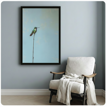 Black framed Scandinavian minimalist hummingbird on a bare twig canvas print with pale blue aged background hung on a soft blue-gray wall beside a white upholstered wood lounge chair with a draped white knit throw
