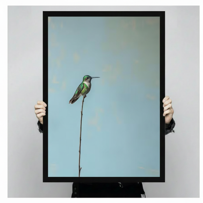 Two hands in black jacket sleeves holding a large black framed minimalist green hummingbird perched on a tall thin branch canvas art print with soft blue background, front-facing vertical product size display