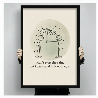 Close-up of hands holding black framed wall art print — stick figures sharing umbrella in rain with heartfelt quote on cream background