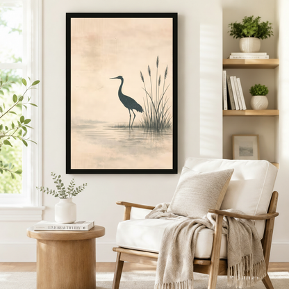 Black framed minimalist Japanese ink wash heron and reed lake canvas art showing a tall dark teal heron standing in a tranquil misty lake with slender reeds and a gentle water reflection on a warm peach distressed parchment background, displayed on a white wall beside a white wooden armchair with a cream throw and a round wood side table with books