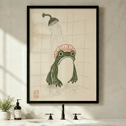 Black framed Japanese woodblock frog shower canvas art with a plump dark green frog wearing a pink ruffled shower cap standing under a vintage rainfall showerhead in a tiled shower with a red seal stamp in the lower left on a warm aged parchment background, displayed above a minimalist bathroom sink with a black faucet, soap dispenser, and small potted plant