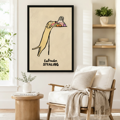 Large black-framed wall art of a mischievous Labrador stealing food in expressive loose sketch style with bold "Labrador Stealing" lettering, styled beside a linen armchair in a Scandinavian-inspired reading nook