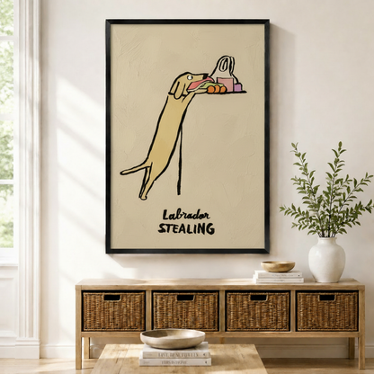 Framed canvas print of a humorous yellow Labrador stretching up to steal food from a counter in loose line art style with "Labrador Stealing" text, displayed above a rattan storage console in a bright modern entryway