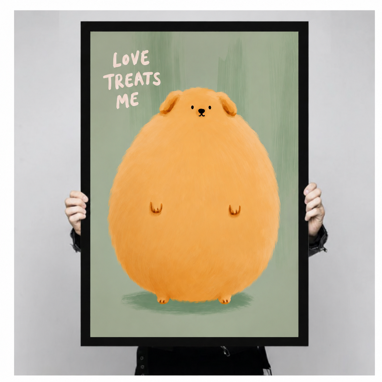 Close-up of a black framed art print featuring a round fluffy cartoon dog with "Love Treats Me" lettering on a soft green background, held up for display
