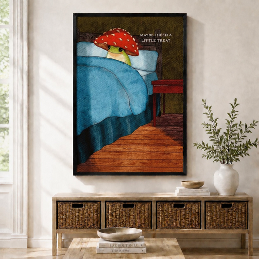 Framed wall art print of a whimsical frog wearing a red mushroom hat tucked in bed with text "Maybe I Need A Little Treat", displayed above a rattan console table in a bright entryway