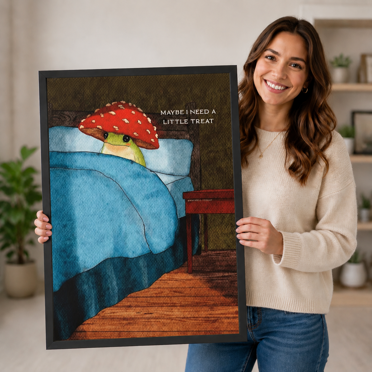 Woman holding a large black-framed art print featuring a green frog in a red mushroom hat tucked into bed, "Maybe I Need A Little Treat" funny quote wall art for home decor