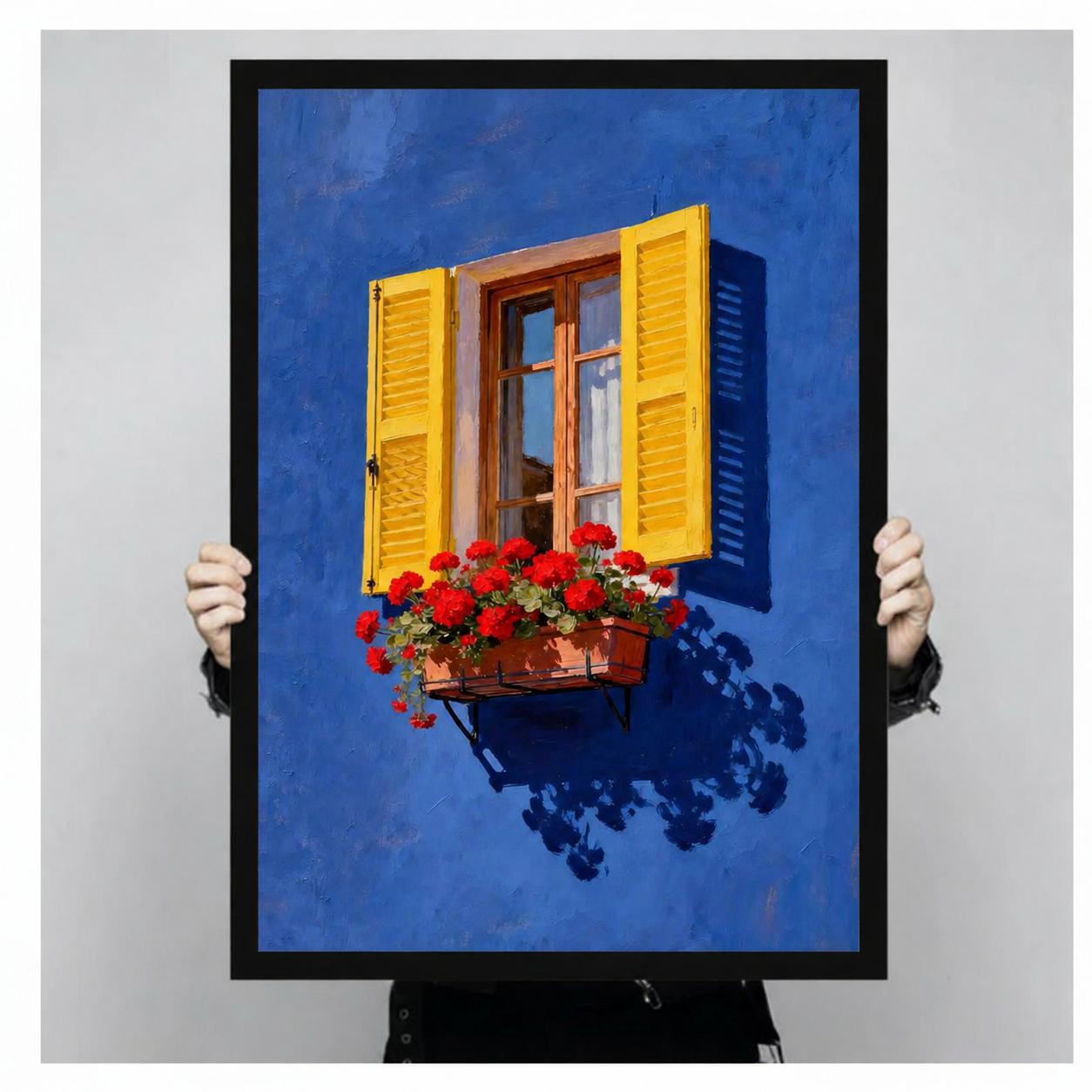 Close-up of black framed wall art print showing Mediterranean blue wall, yellow louvered shutters and red geranium flower box held by person