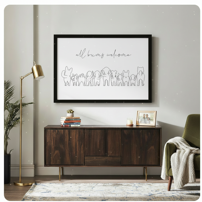 Black framed minimalist all bums welcome line art canvas art showing flowing script lettering above a humorous lineup of animal back view outlines including a rabbit, cat, dogs, elephant, pig, and French bulldog in fine single-stroke black line art on a white background, hung above a dark walnut wood console cabinet with a gold floor lamp, potted palm, olive green armchair with a cream knit throw, stacked books, and a framed photo