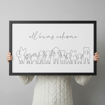 Person in a cream cable-knit sweater holding a large horizontal black framed minimalist all bums welcome line art canvas art print showing elegant script lettering above a playful row of animal back view silhouettes including a rabbit, cat, dogs, elephant, pig, and French bulldog in fine single-stroke black line art on a white background against a light gray backdrop