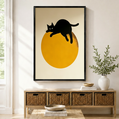Black framed mid-century minimalist black cat moon canvas art with a sleek black cat silhouette lazily draped over the top of a large bold golden orange circle with legs dangling and tail curled upward and a cool piercing gaze on a clean warm off-white background in a striking three-color black gold and cream palette, displayed above a wooden rattan storage bench with a white ceramic bowl, stacked books, and a white vase of greenery