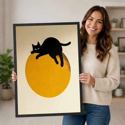 Smiling woman in a cream sweater holding a large vertical black framed mid-century minimalist black cat moon canvas art print showing a sleek black cat silhouette lazily sprawled over a large saturated golden orange circle with legs hanging loose and tail curled upward on a warm off-white background in a bold three-color black amber and cream palette in a bright plant-filled room