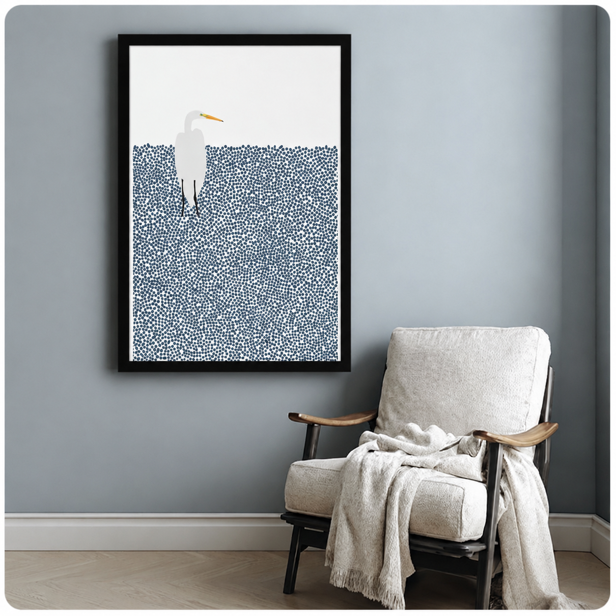 Black framed minimalist white egret with yellow beak standing in dotted navy blue water art print on a blue-grey wall beside a white linen armchair with draped throw