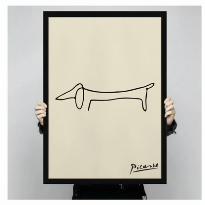 Person holding a black framed Picasso minimalist one-line dachshund sketch art print with handwritten signature on cream background against a light gray backdrop