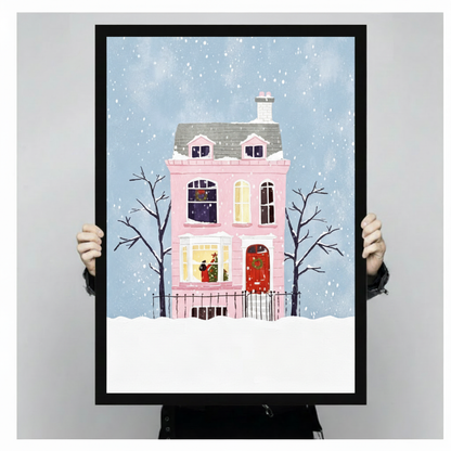 Whimsical flat illustration of a pink Victorian house with a red door and wreath surrounded by bare trees in falling snow Christmas wall art framed print held against light gray background