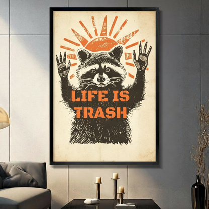Black framed retro linocut raccoon with arms raised and an exasperated expression beneath an orange woodcut sun with bold Life Is Trash stencil text humor poster canvas wall art on aged cream background displayed in a modern gray living room with dark sofa and metal candlestick holders