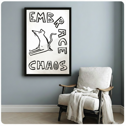 Black framed witty minimalist rat illustration with thick black outline and expressive chaotic pose beside bold Embrace Chaos hand-drawn typography humor canvas print on white background hung on a soft blue-gray wall beside a white linen wood folding chair with a draped cream throw