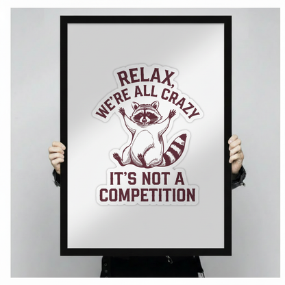 Person holding a black framed art print featuring a raccoon illustration and the humorous quote Relax We're All Crazy It's Not A Competition against a neutral gray background