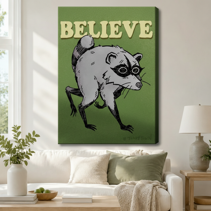 Stretched canvas retro funny Believe raccoon wall art featuring a sketchy gray raccoon with a black eye mask in a low crouching stance with bold cream Believe lettering at the top on a deep olive green background, hung above a white sofa with sage green pillows in a bright living room