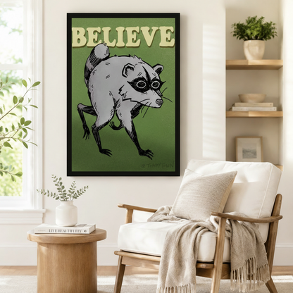 Black framed retro funny Believe raccoon canvas art showing a scruffy gray raccoon with a black mask crouching with clawed feet and a grumpy expression under bold cream Believe text on a rich olive green background, displayed beside a white linen armchair in a cozy reading nook