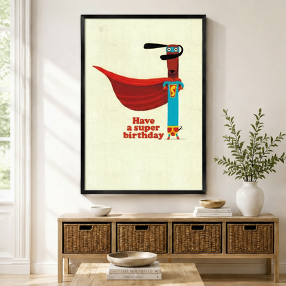 Black framed retro funny superhero dachshund Have a Super Birthday canvas art with a slim brown dachshund in a teal superhero suit with a bold red cape and blue mask on a vintage cream background, displayed above a rattan storage console in a bright entryway
