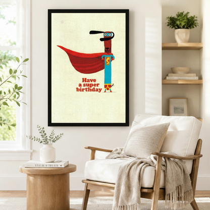 Black framed retro funny superhero dachshund Have a Super Birthday canvas art showing a slim brown dachshund in a teal superhero suit with a dramatic red cape and blue eye mask on a vintage cream background, displayed beside a white linen armchair in a cozy reading nook