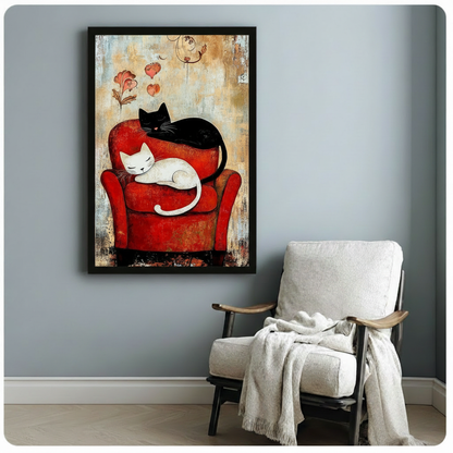 Black framed vintage-style black and white cat love canvas art with a drowsy white cat resting in a rich red armchair and a black cat draped over the top, with floating hearts and a hand-drawn flower on a warm crackled background, displayed on a blue-gray wall beside a white linen folding chair with a cream throw