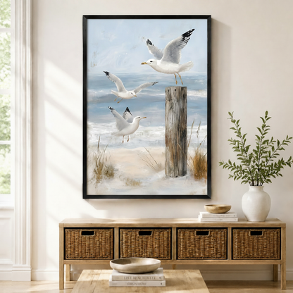 Framed seagull beach wall art print featuring three white gulls in flight over sandy dunes and a weathered wooden post with soft blue ocean waves, displayed above a wicker console in a bright entryway