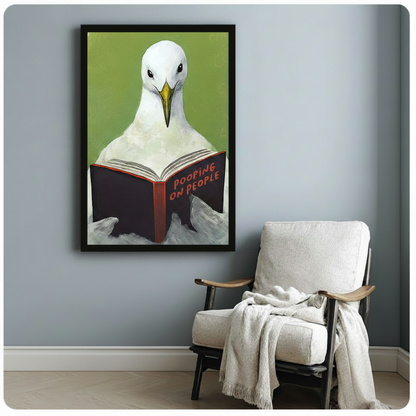 Black framed humorous coastal seagull reading a Pooping on People book canvas print with bold olive green background hung on a soft blue-gray wall beside a white linen wood lounge chair with a draped cream knit throw