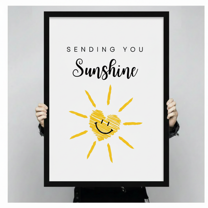 Hands holding a large black framed canvas print of a yellow smiley sun with a cross-hatched heart face and Sending You Sunshine quote in bold script, cheerful hand-drawn sunshine illustration on white background against a light gray backdrop