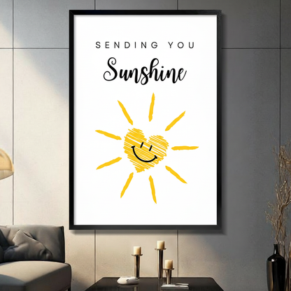 Black framed canvas wall art with Sending You Sunshine quote in mixed serif and script typography above a hand-drawn yellow sun with a heart-shaped smiley face, cheerful doodle illustration on white background, displayed above a dark console table with three pillar candles and a black vase against a gray slate paneled wall