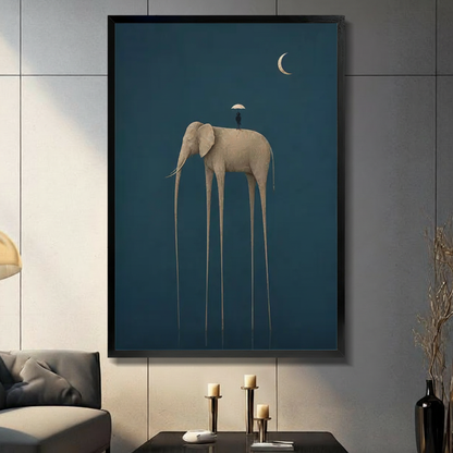 Black framed surrealist elephant canvas art print featuring a long-legged golden elephant with a tiny umbrella figure on its back and a crescent moon, displayed on a grey wall above a console table with gold candle holders in a modern living room