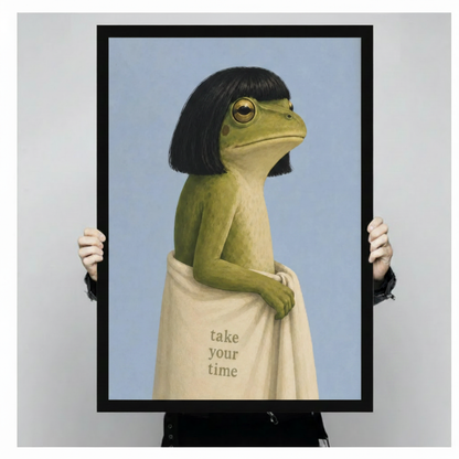 Person holding a large black framed surreal green frog canvas art print featuring a stoic frog with black bob hair wrapped in a cream towel with take your time lettering against a light gray studio background