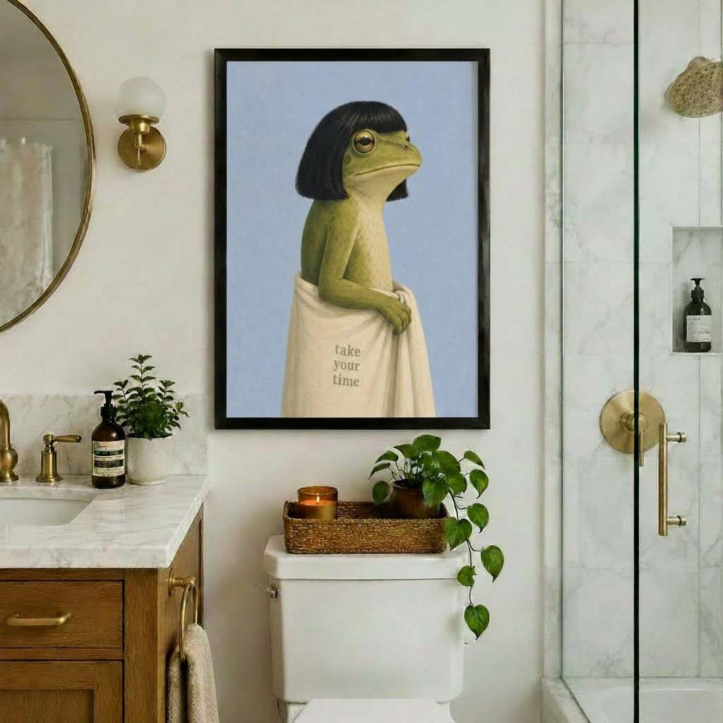 Black framed whimsical frog with bob haircut and cream take your time towel canvas art hung above a toilet in an elegant bathroom with brass fixtures, marble vanity, round mirror, and glass shower enclosure
