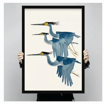 Hands holding large framed three flying herons canvas print - full artwork detail of Japanese ukiyo-e watercolor painting with three graceful herons in layered flight formation blue grey gradient feathers bold yellow eyes black crest plumes and golden talon legs on clean cream background