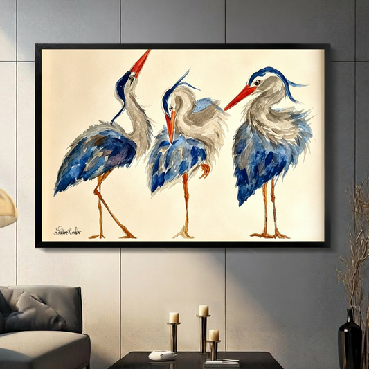 Whimsical watercolor illustration of three blue-gray herons in playful poses a tall neck-stretching bird a plump hunched bird and a head-tilting bird with red-orange beaks and orange legs on a cream background witty heron wall art framed print in a living room with dark gray panel wall dark sofa and black vase