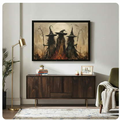 Black framed dramatic gothic Halloween oil painting of three witches in flowing black robes and tall pointed hats standing back-to-viewer under a large full moon with bats flying gnarled staffs in hand and a bonfire glowing at their feet warm amber and dark brown toned canvas wall art hung above a dark wood sideboard with stacked books succulents and a gold floor lamp beside an olive green armchair with cream throw