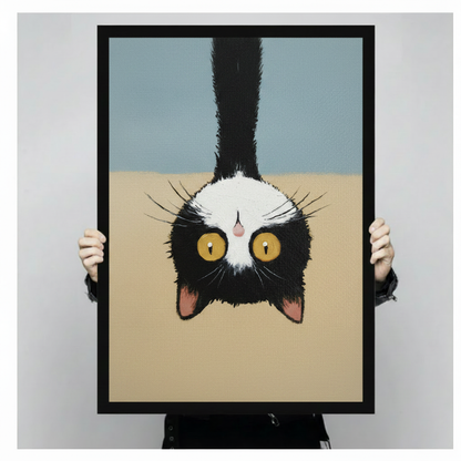 Hand-held framed print of a tuxedo cat hanging upside down, full artwork view showing wide golden eyes and fluffy black and white fur against blue and sandy beige background