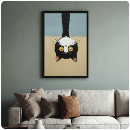 Humorous tuxedo cat portrait hanging head-down with bold golden eyes against minimalist blue-beige background, black framed print above beige fabric sofa