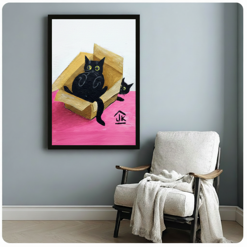Funny black cats cardboard box wall art, chubby cat lounging inside box while small cat peeks from outside, black framed print above white linen armchair with draped blanket