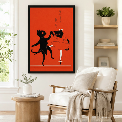 Large black framed retro Japanese dancing cat poster in red and black beside a white linen armchair, round wooden side table with stacked books, and potted greenery by a sunny window