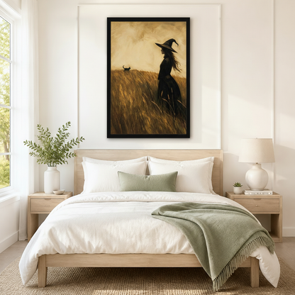 Black framed vintage folk art witch and black cat in a wheat field canvas art with a long-haired witch in a pointed hat standing in a golden field beside a small black cat walking through the grass in warm sepia oil-paint texture, hung above a wooden bed frame in a sun-drenched bedroom with crisp white linen bedding, a sage green throw, and a round white nightstand lamp