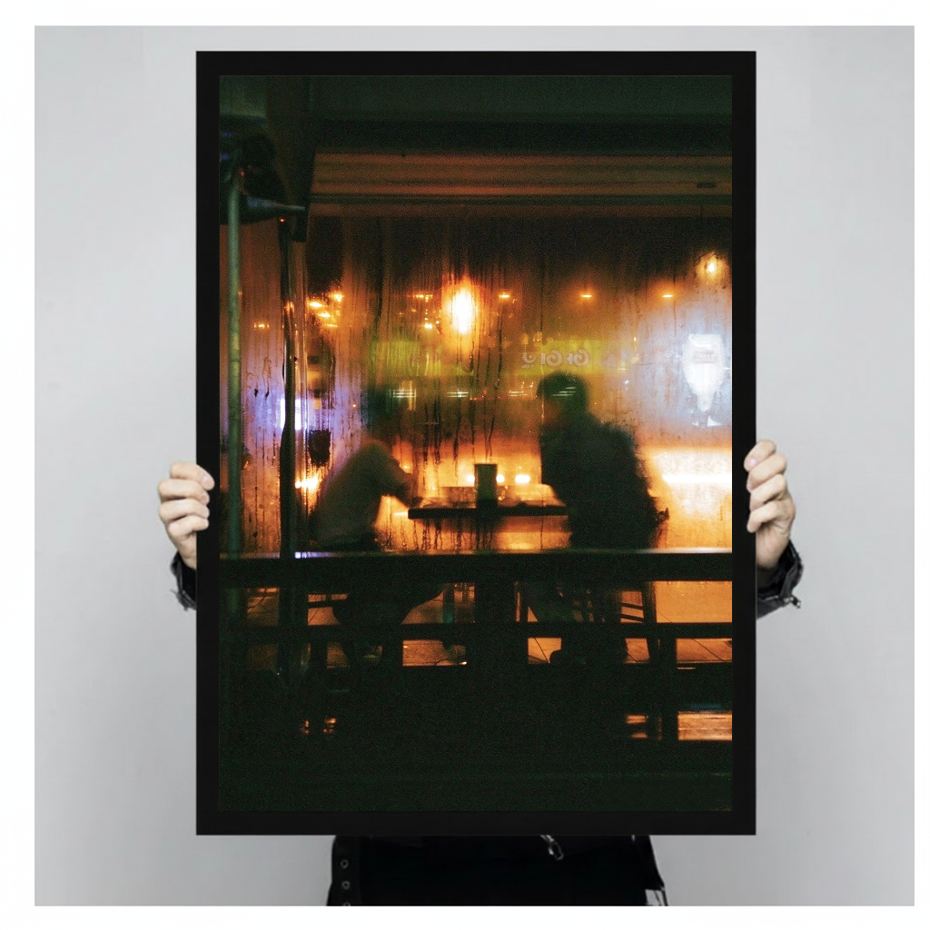 Warm cafe silhouettes behind a rainy window in black framed canvas held by a person.