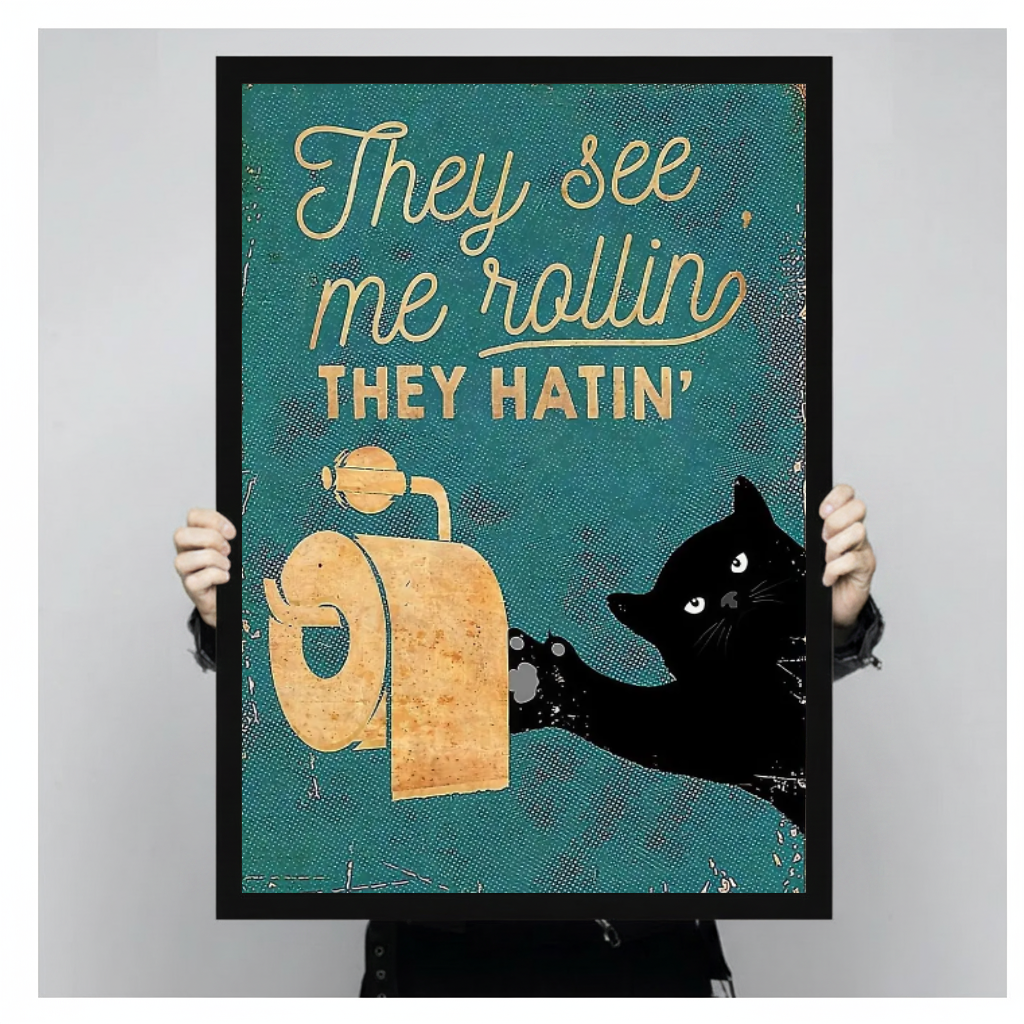 Person holding black framed teal toilet paper cat artwork against a plain pale background.